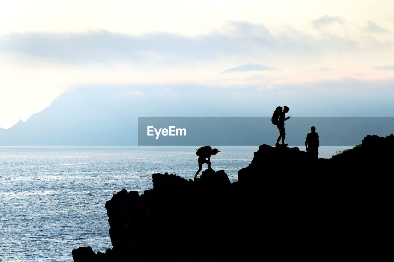 Silhouette hikers standing on mountain by sea against cloudy sky