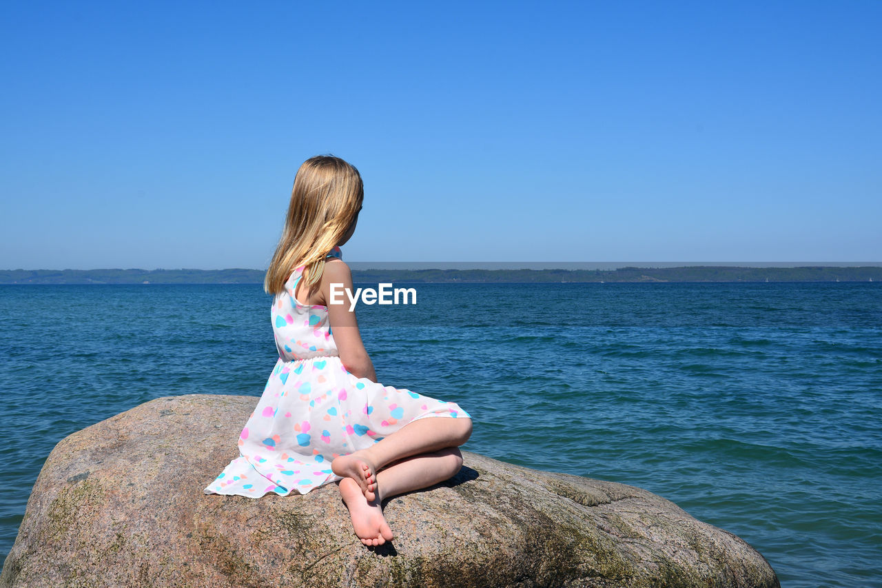 Side view of girl sitting on rock by sea against clear sky