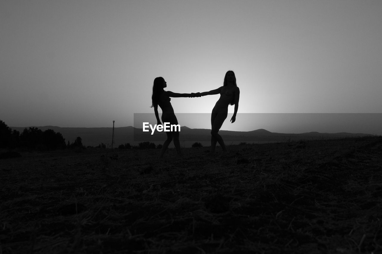 Silhouette women holding hands on landscape against sky during sunset