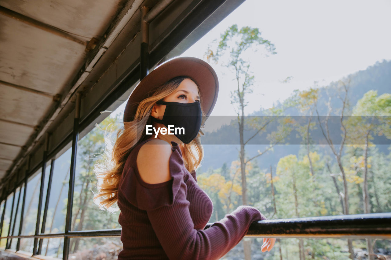 Low angle view of woman standing wearing a mask looking outdoors through a window