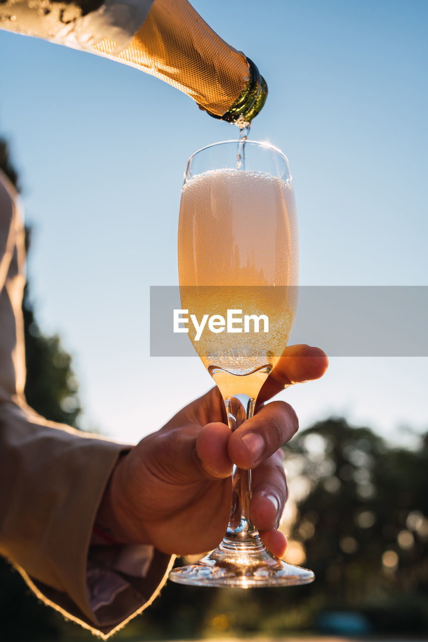 Crop anonymous person having champagne poured into glass from bottle on blurred background of outdoor poolside in evening