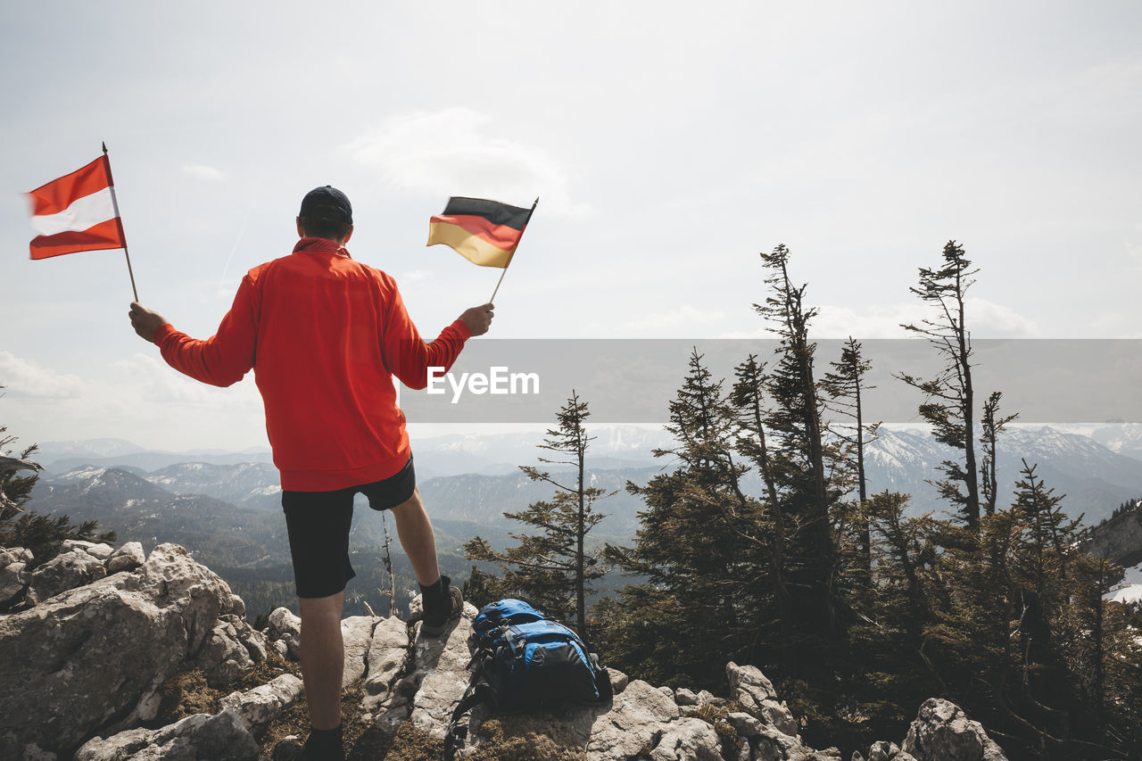 Rear view of mature man holding national flags while standing on mountain against sky