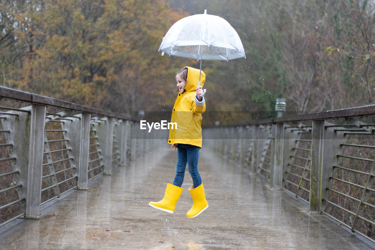 Full body side view of cheerful little girl in bright yellow raincoat and rubber boots with umbrella in hand jumping on wet bridge in rainy autumn weather in park