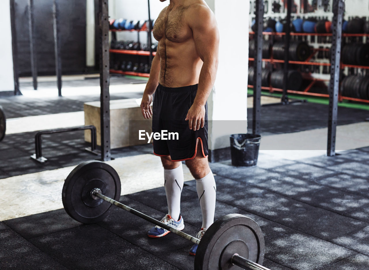 Low section of shirtless man standing by barbell in gym