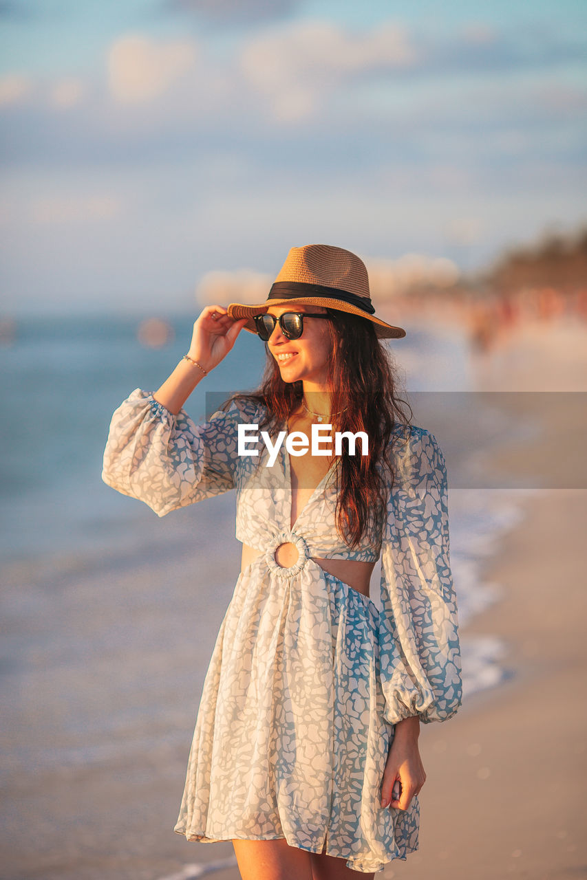 one person, clothing, beach, women, adult, sea, water, land, fashion, hat, young adult, nature, sky, spring, vacation, trip, long hair, holiday, summer, standing, hairstyle, sunset, blue, dress, leisure activity, sun hat, smiling, casual clothing, photo shoot, travel, happiness, three quarter length, beauty in nature, sand, portrait, emotion, female, relaxation, travel destinations, lifestyles, sunglasses, looking, brown hair, glasses, sunlight, tourism, outdoors, copy space, front view, person, focus on foreground, carefree, enjoyment, day, cheerful, tourist, positive emotion, cloud, fashion accessory, sun, fun, fedora