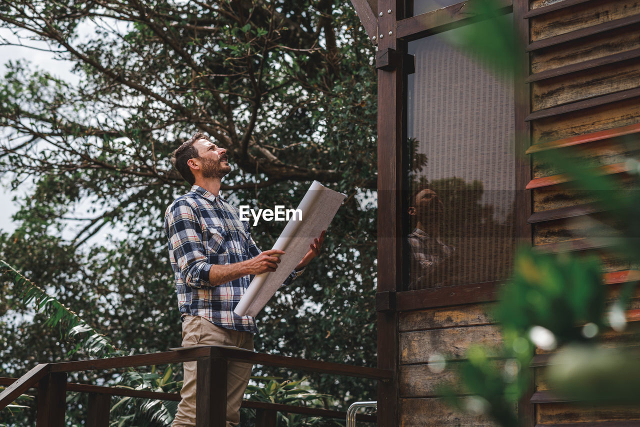 Low angle side view of male architect with paper draft standing on terrace of wooden house in forest