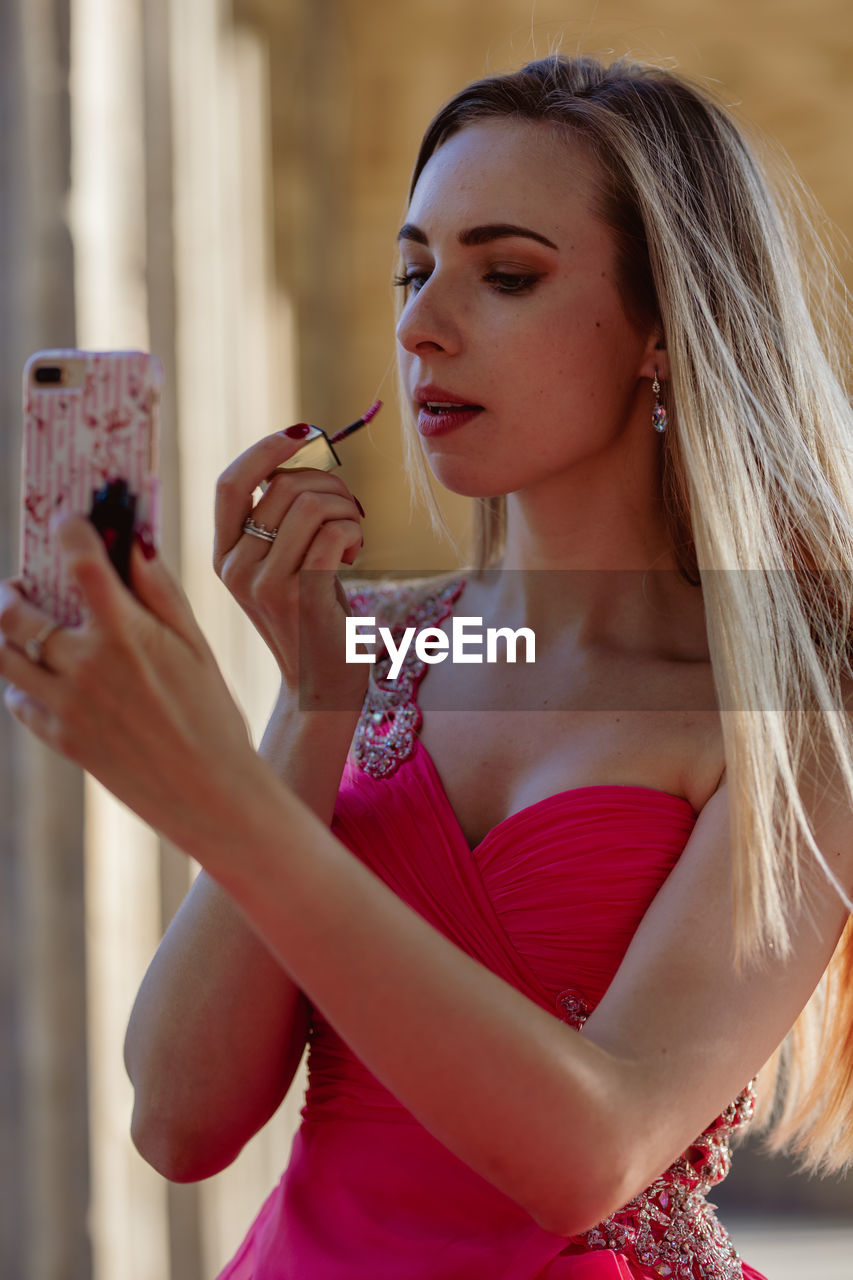 Young woman in pink evening gown applying lipstick while looking at smart phone outdoors