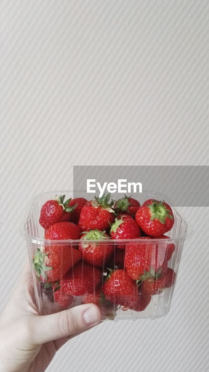 Cropped hand holding strawberry in container against wall