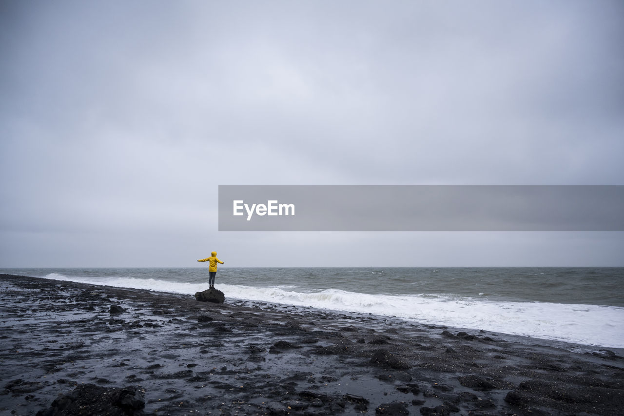 Woman with arms outstretched standing on rock at sea shore against cloudy sky