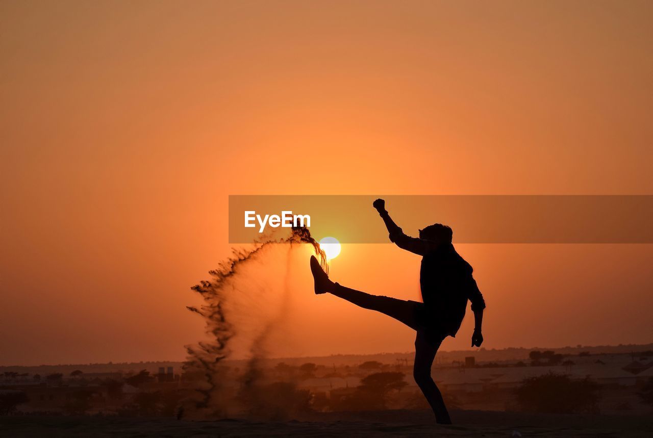 Silhouette man with arms raised playing with sand against sky during sunset