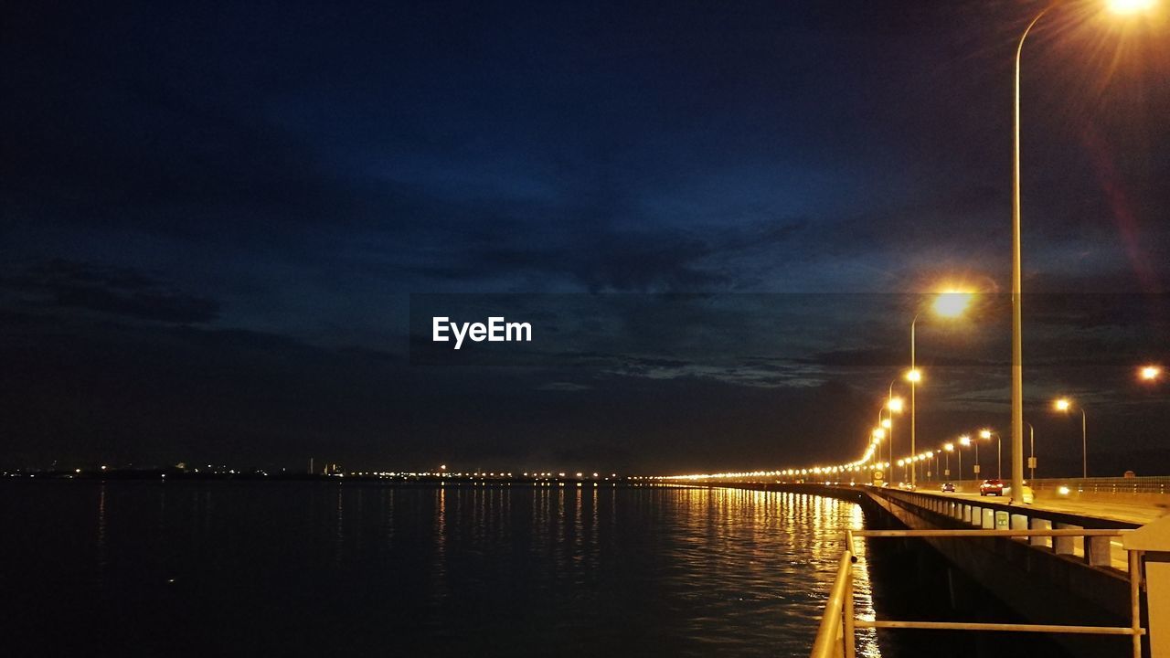 ILLUMINATED BRIDGE OVER CALM SEA AGAINST SKY