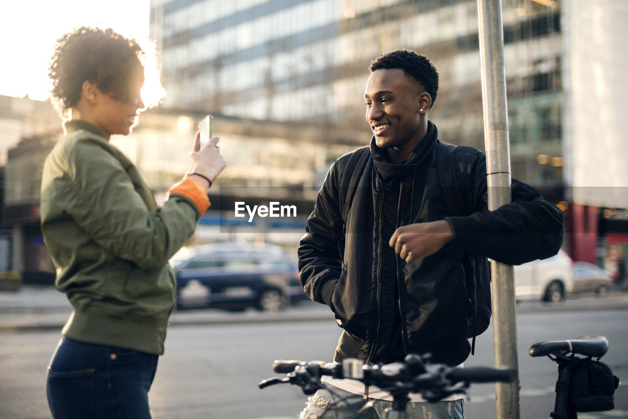 Side view of teenager photographing friend through smart phone while standing by city street