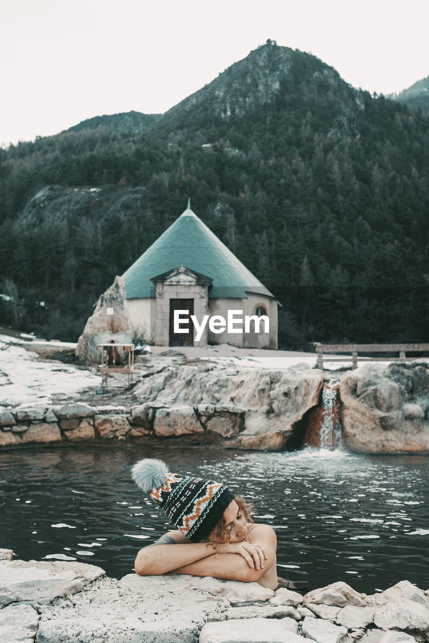 rear view of woman sitting on rock by lake against mountain