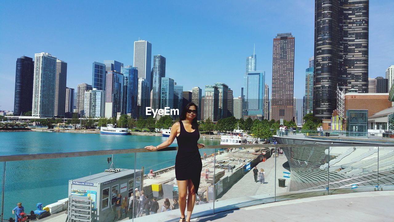 Portrait of young woman standing against modern buildings and river in city