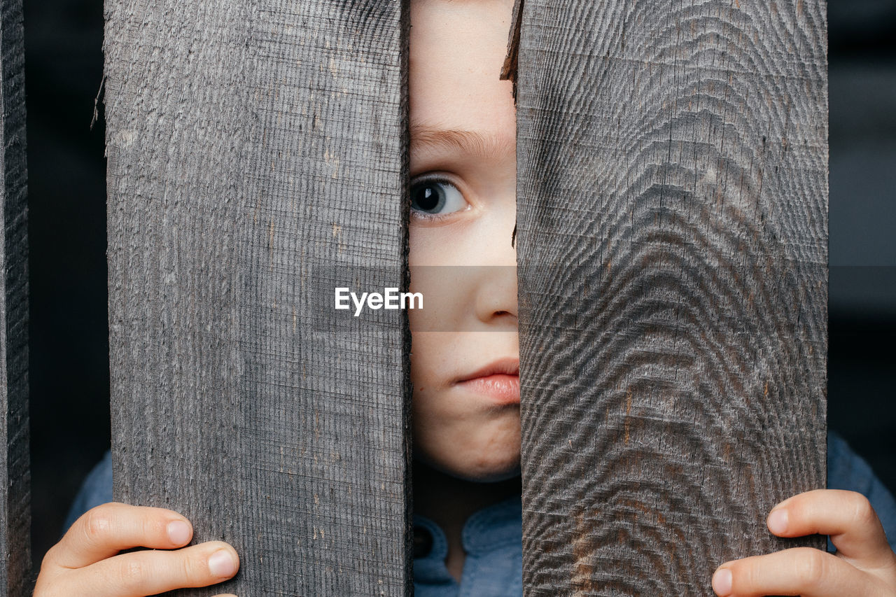 Close-up of a young male guy looking out from behind a rustic wooden fence, looking into the camera