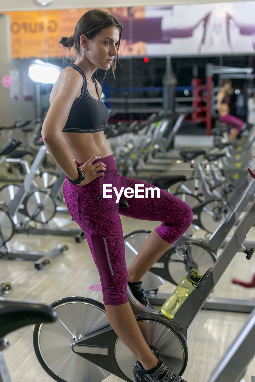 Concentrated sweaty young sportswoman in top and legging doing cardio training on exercise bike in gym
