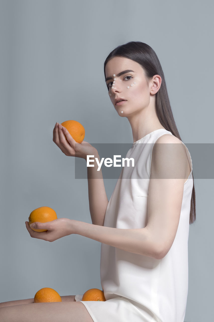 Conceptual studio portrait of a young female model holding orange fruit on light blue backgroung