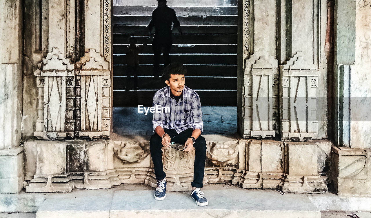 Full length of teenage boy looking away while sitting on entrance