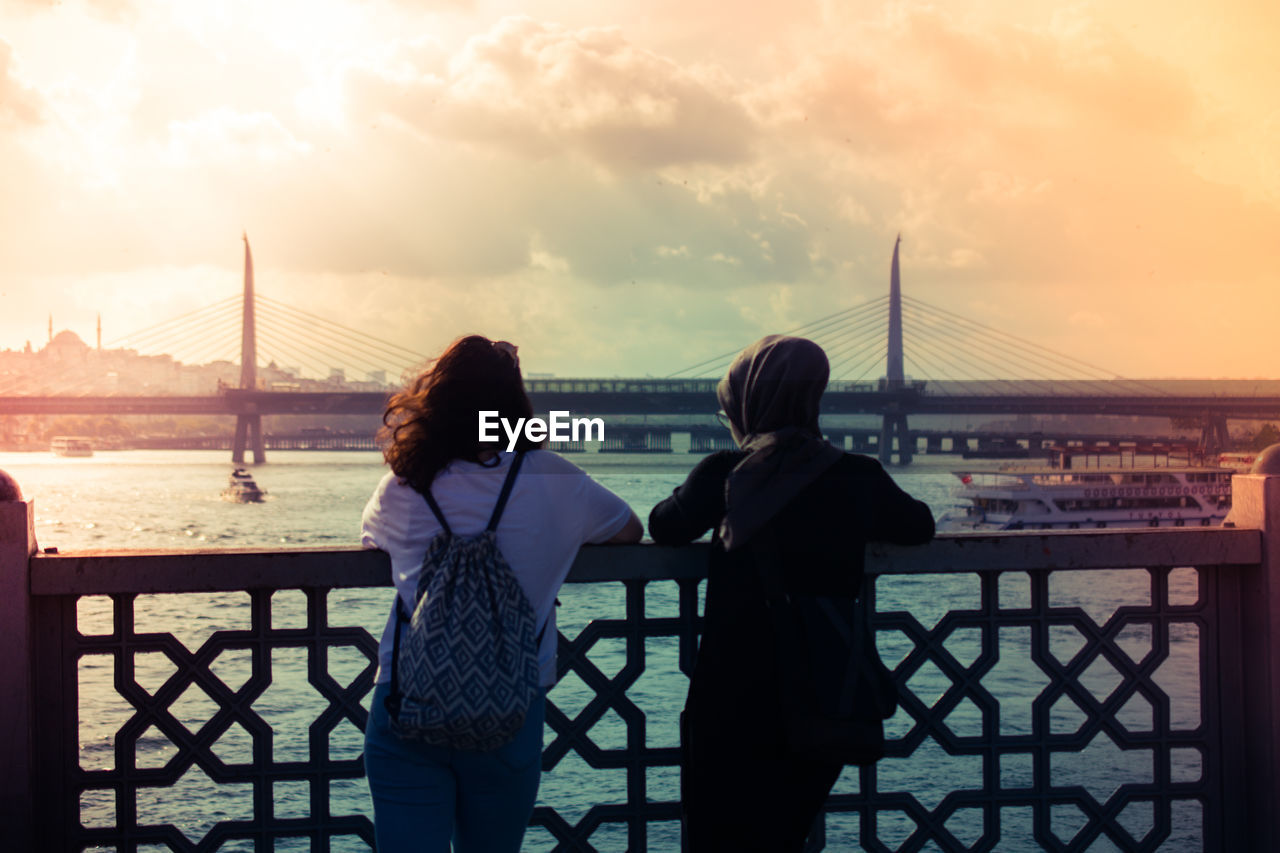 REAR VIEW OF WOMEN STANDING ON BRIDGE OVER SEA AGAINST SKY