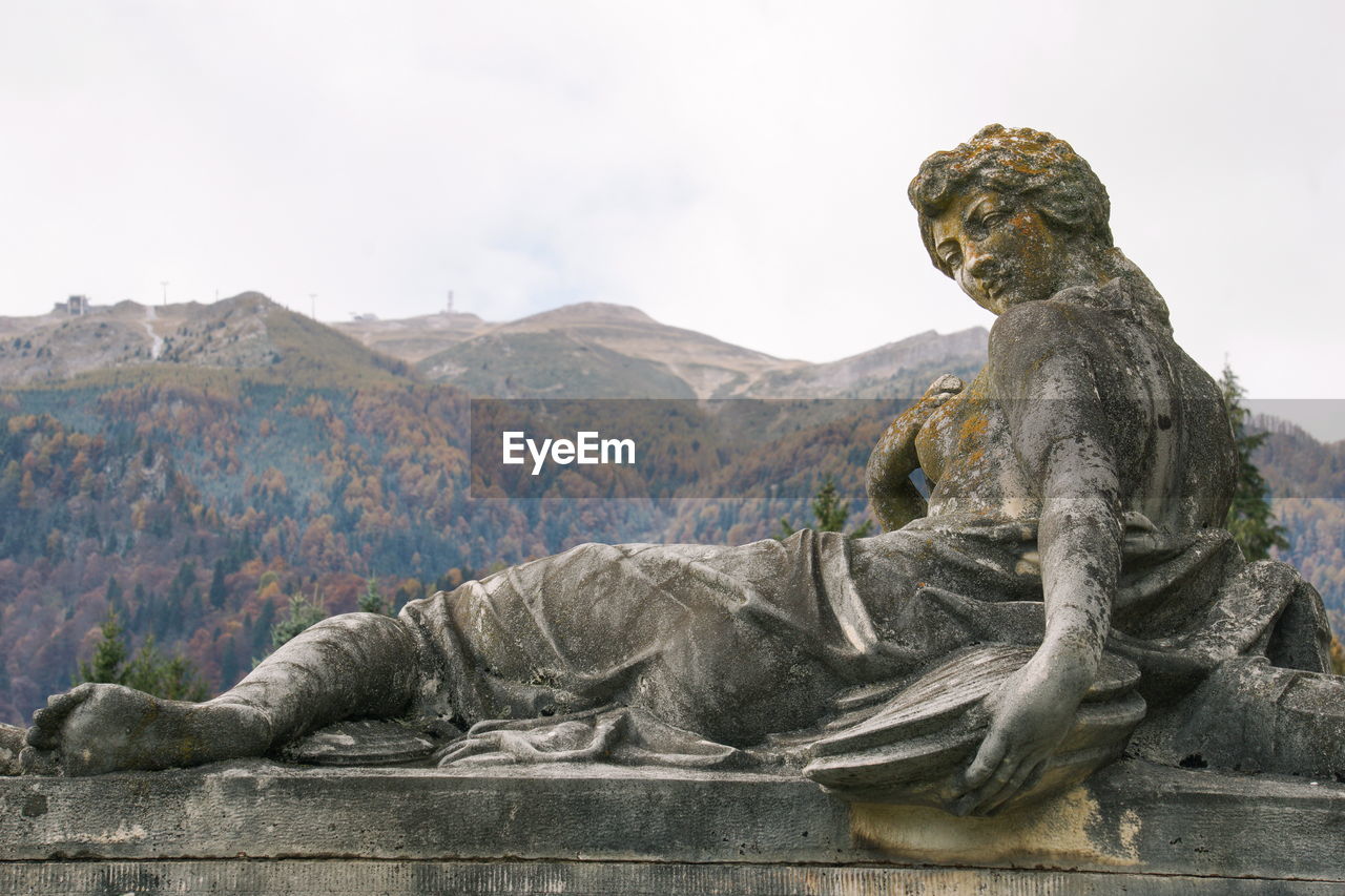 Close-up of old weathered female statue by mountains against sky