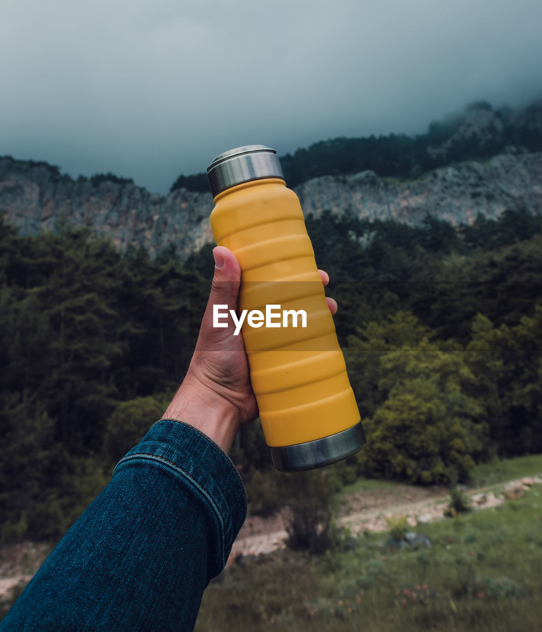 Male hand holding a steel yellow thermo bottle on nature background.