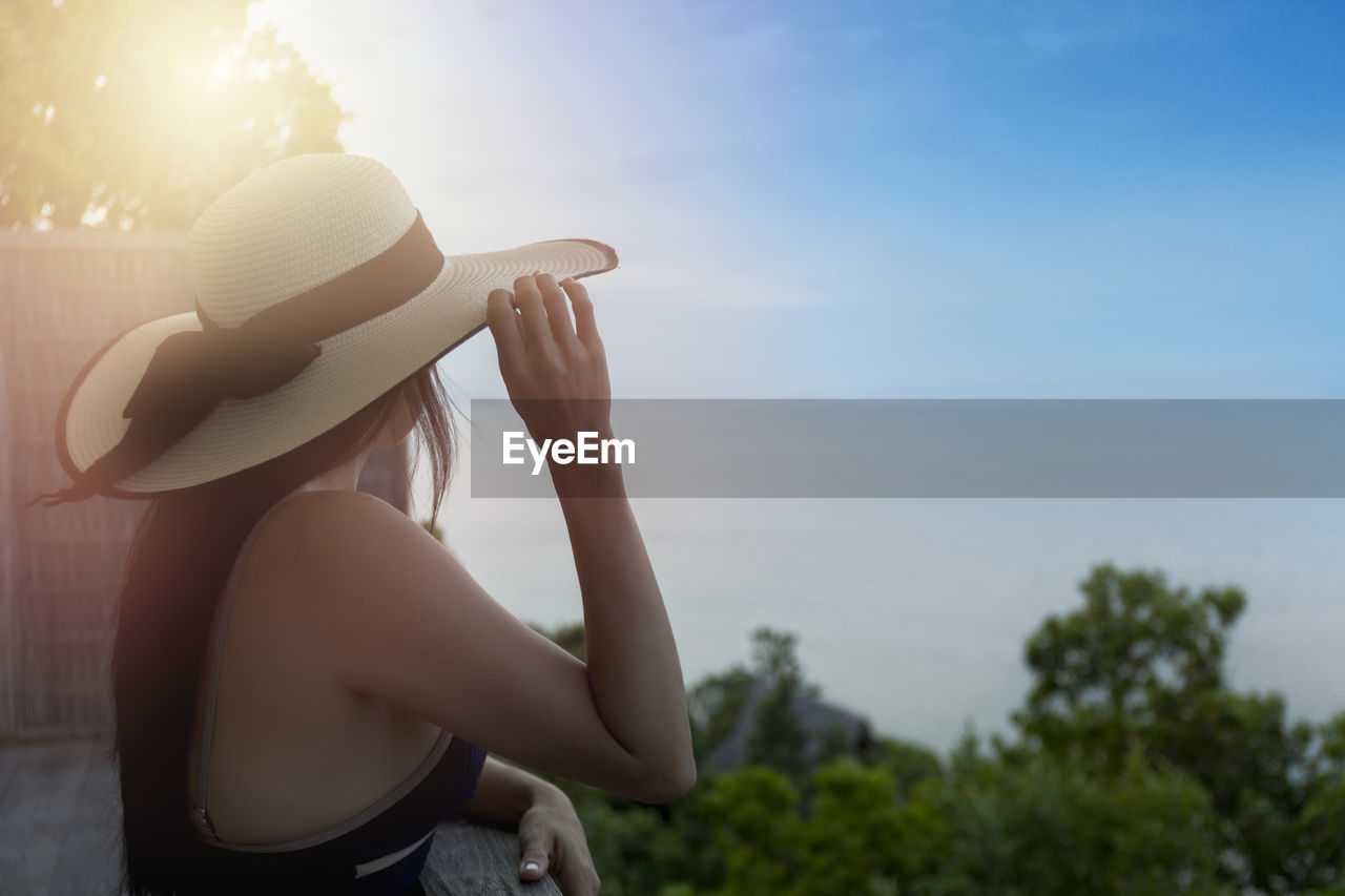 one person, sky, adult, nature, hat, sunlight, women, summer, clothing, sun hat, young adult, sun, vacation, trip, copy space, land, lens flare, holiday, relaxation, day, sunset, water, beauty in nature, fashion accessory, outdoors, leisure activity, fedora, lifestyles, plant, tranquility, travel, side view, person, female, back lit, sea, fashion, hand, tree, hairstyle, landscape, holding, beach, tranquil scene, waist up, long hair, casual clothing