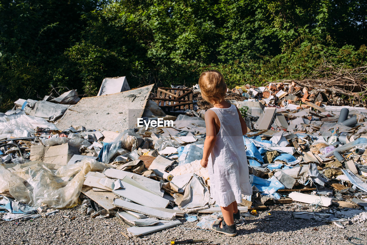 Little girl at a dump among a heap of scattered garbage in the forest.