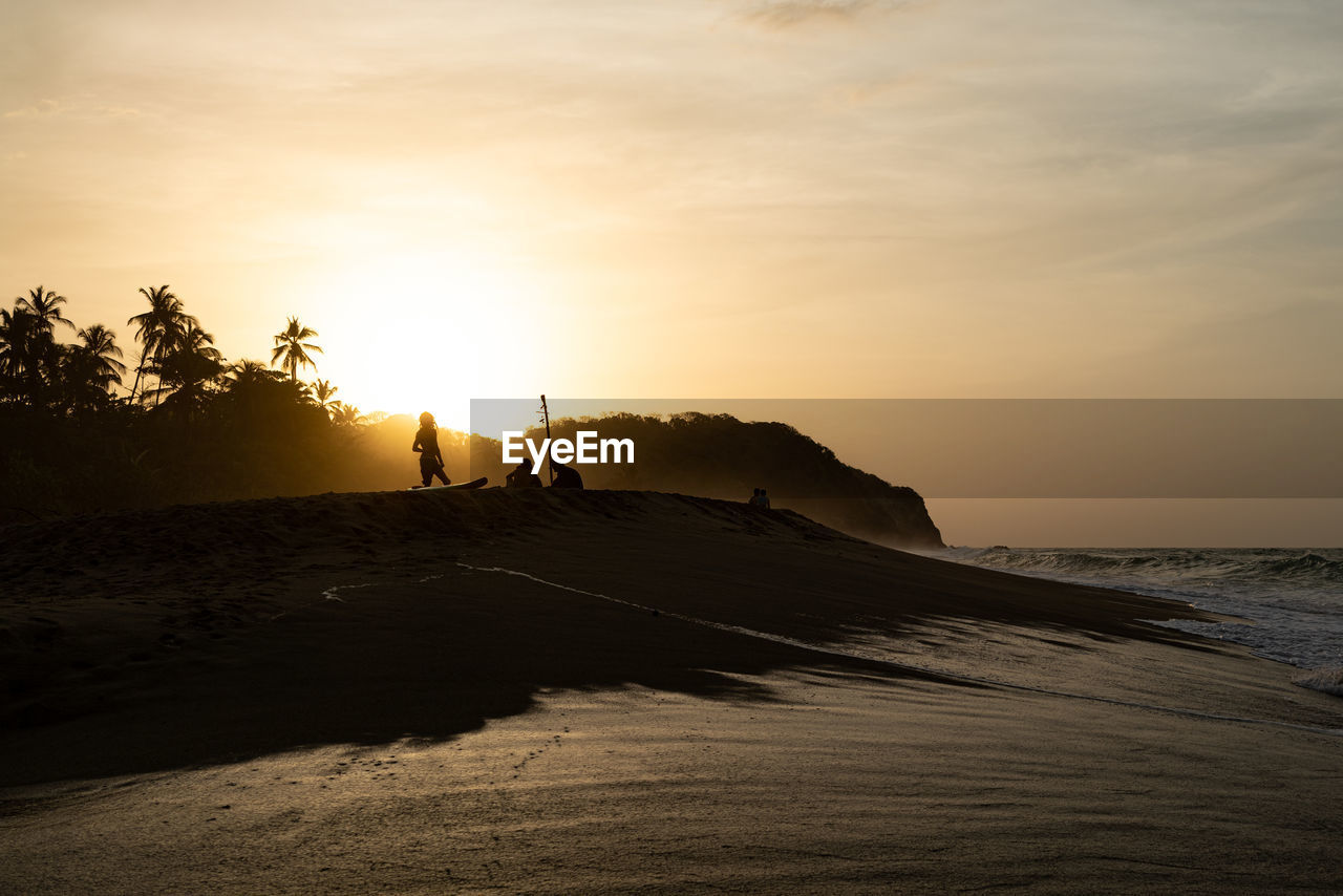 Scenic view of beach and surfers against sky during sunset