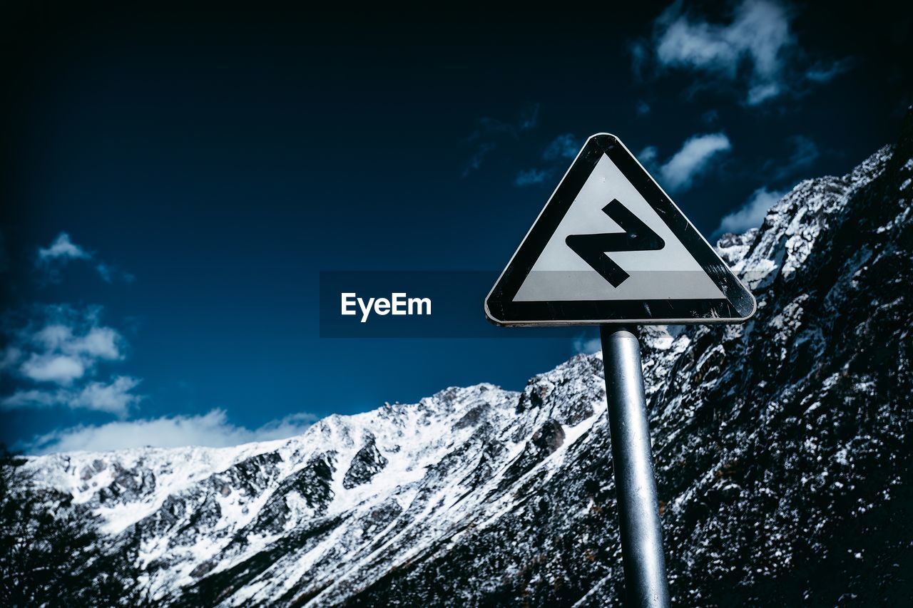 sign, communication, guidance, road sign, sky, road, snow, nature, mountain, no people, warning sign, cold temperature, symbol, winter, cloud, scenics - nature, arrow symbol, directional sign, blue, environment, information sign, outdoors, beauty in nature, darkness, day, triangle shape, transportation, non-urban scene, white, landscape, light, low angle view, mountain range