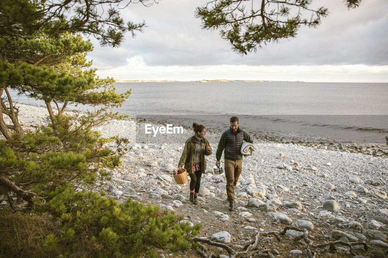 Couple carrying basket and blanket at beach against sky during sunset