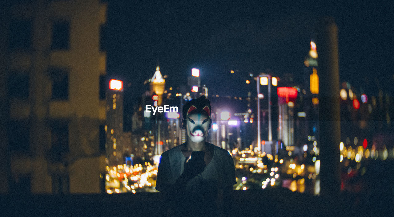 Man wearing mask while using mobile phone against cityscape at night