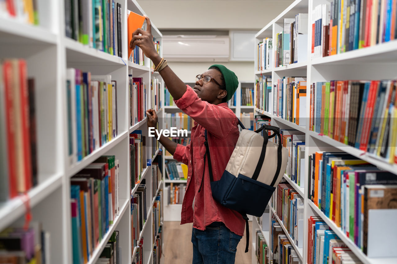 Nerd student african american man choosing book in university library taking it from shelf.