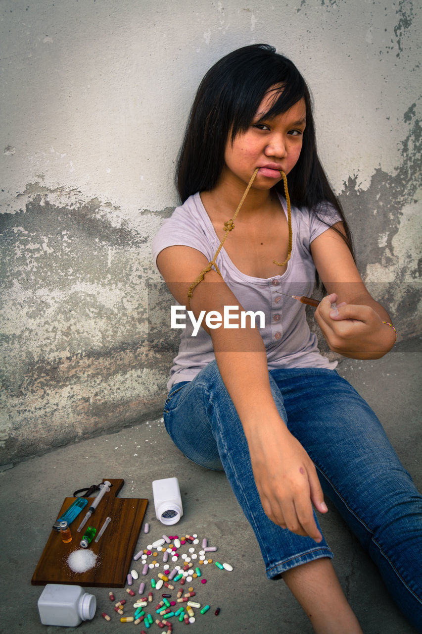 Sad teenage girl with drugs sitting on floor against wall