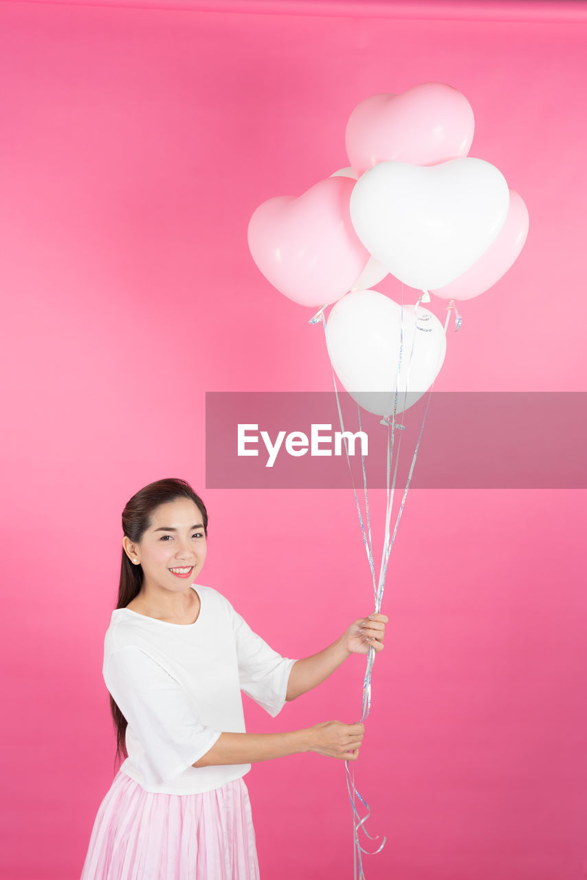 Portrait of happy woman holding heart shape helium balloons while standing against pink background