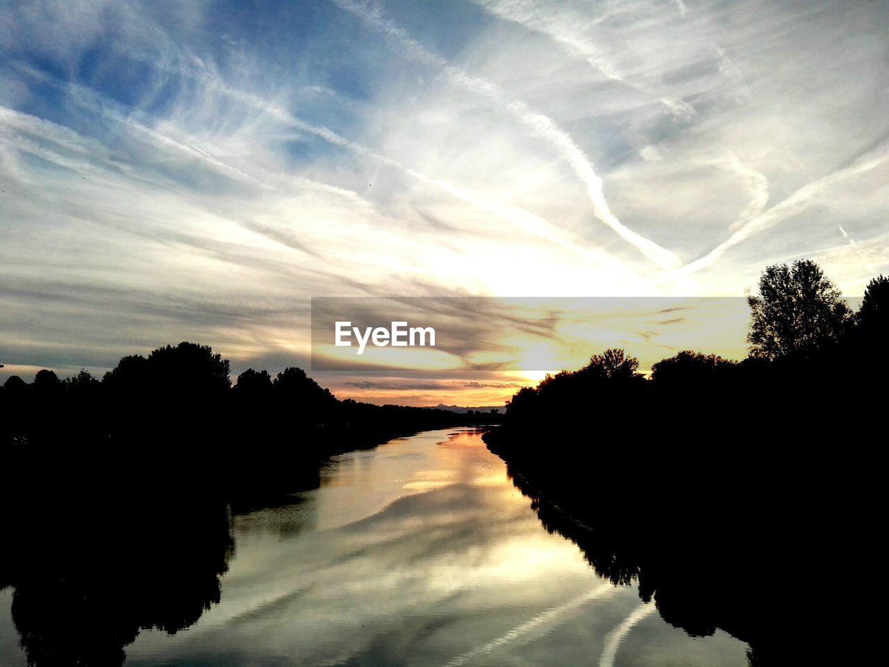 Scenic view of river amidst silhouette trees against cloudy sky at sunset