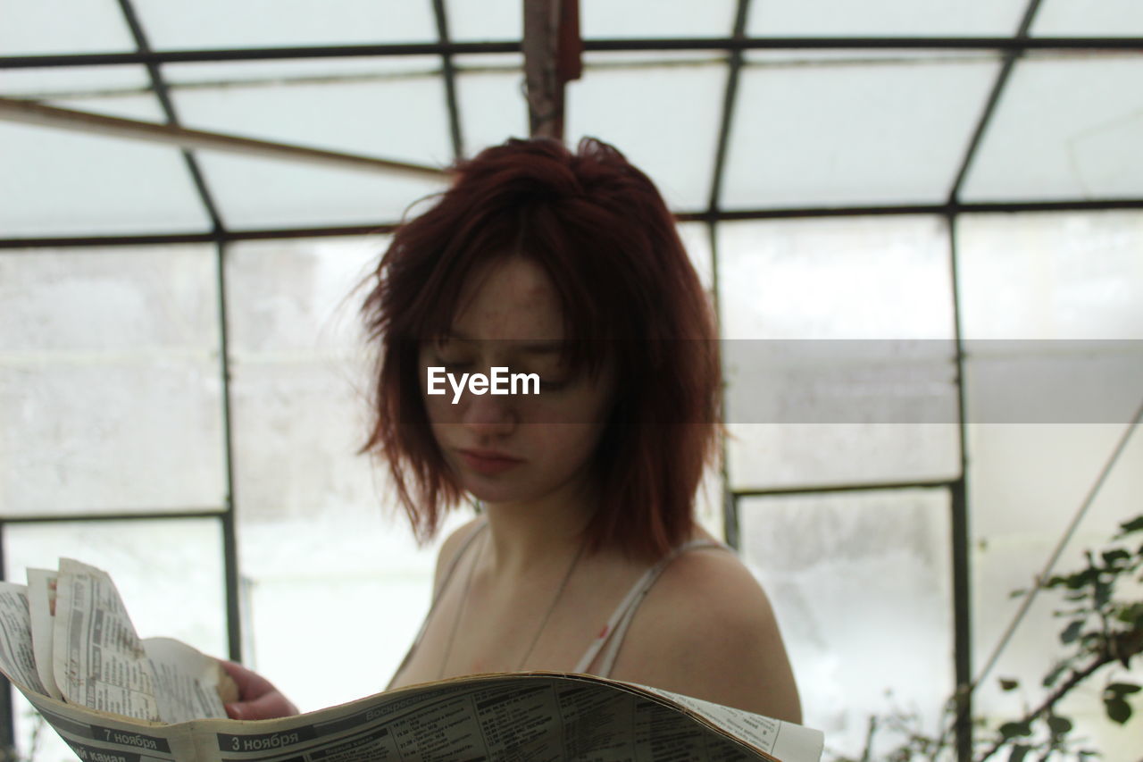 Young woman reading newspaper against window