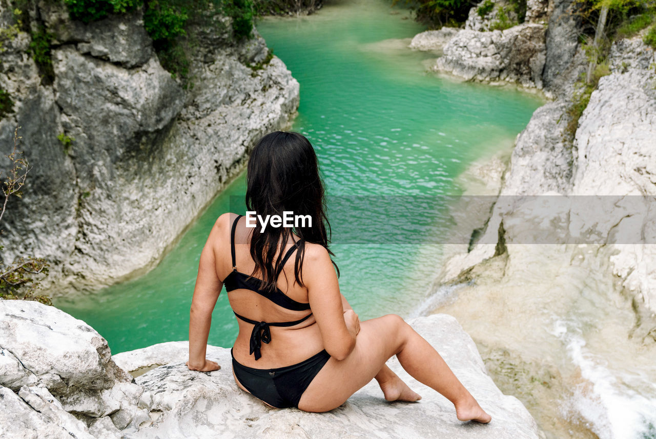 Young woman in bikini sitting on rock by river, waterfall. bikini, outdoors, beautiful girl.