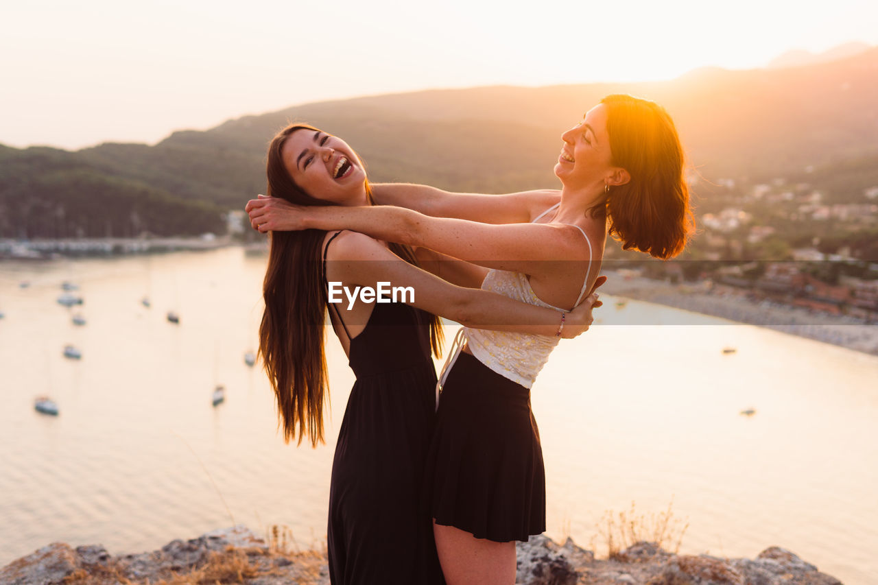 Two women laugh and embrace as friends on a cliffside overlooking a bay with boats at golden hour.