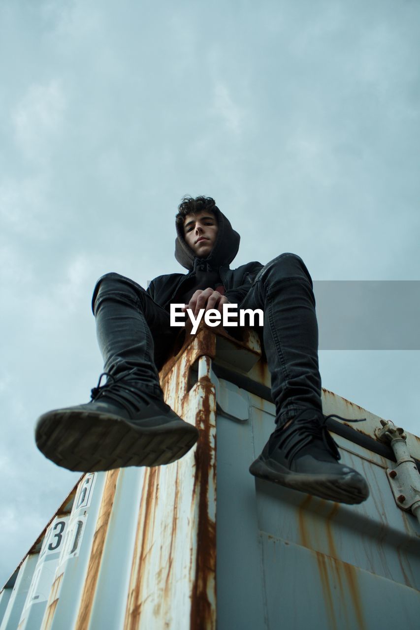 Low angle portrait of teenage boy sitting on metal structure against cloudy sky