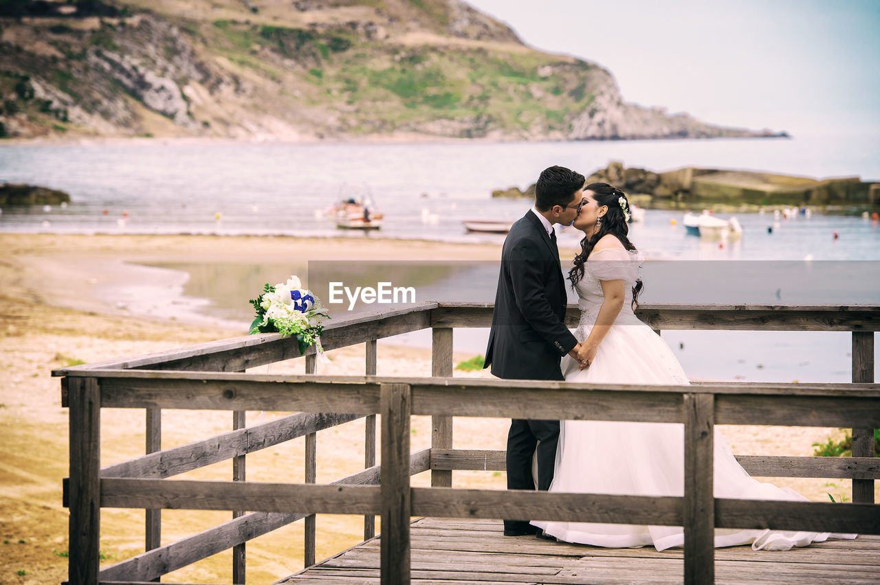 Couple kissing while standing on pier by sea against sky