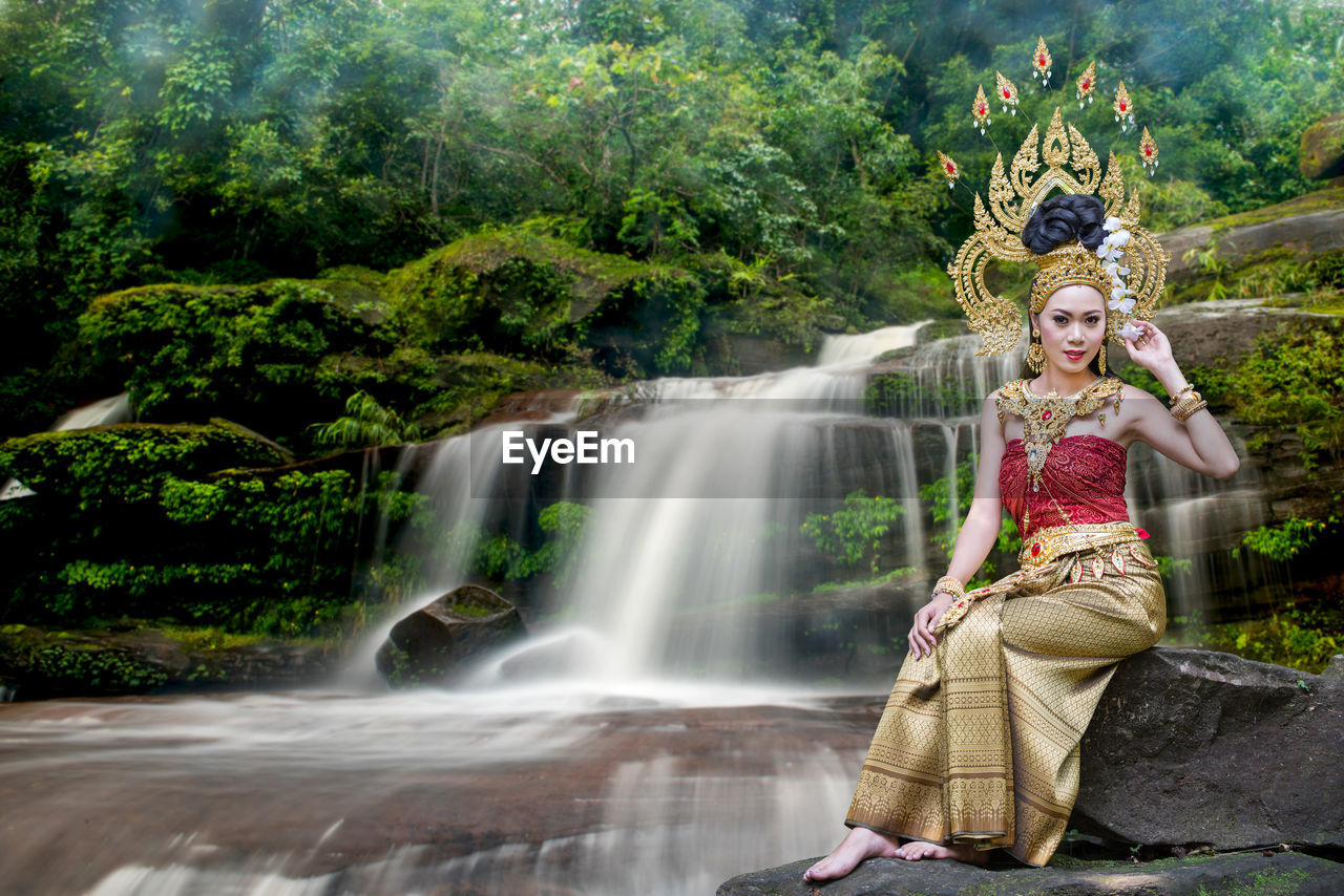 Portrait of beautiful young woman wearing crown and traditional clothing while sitting against waterfall in forest