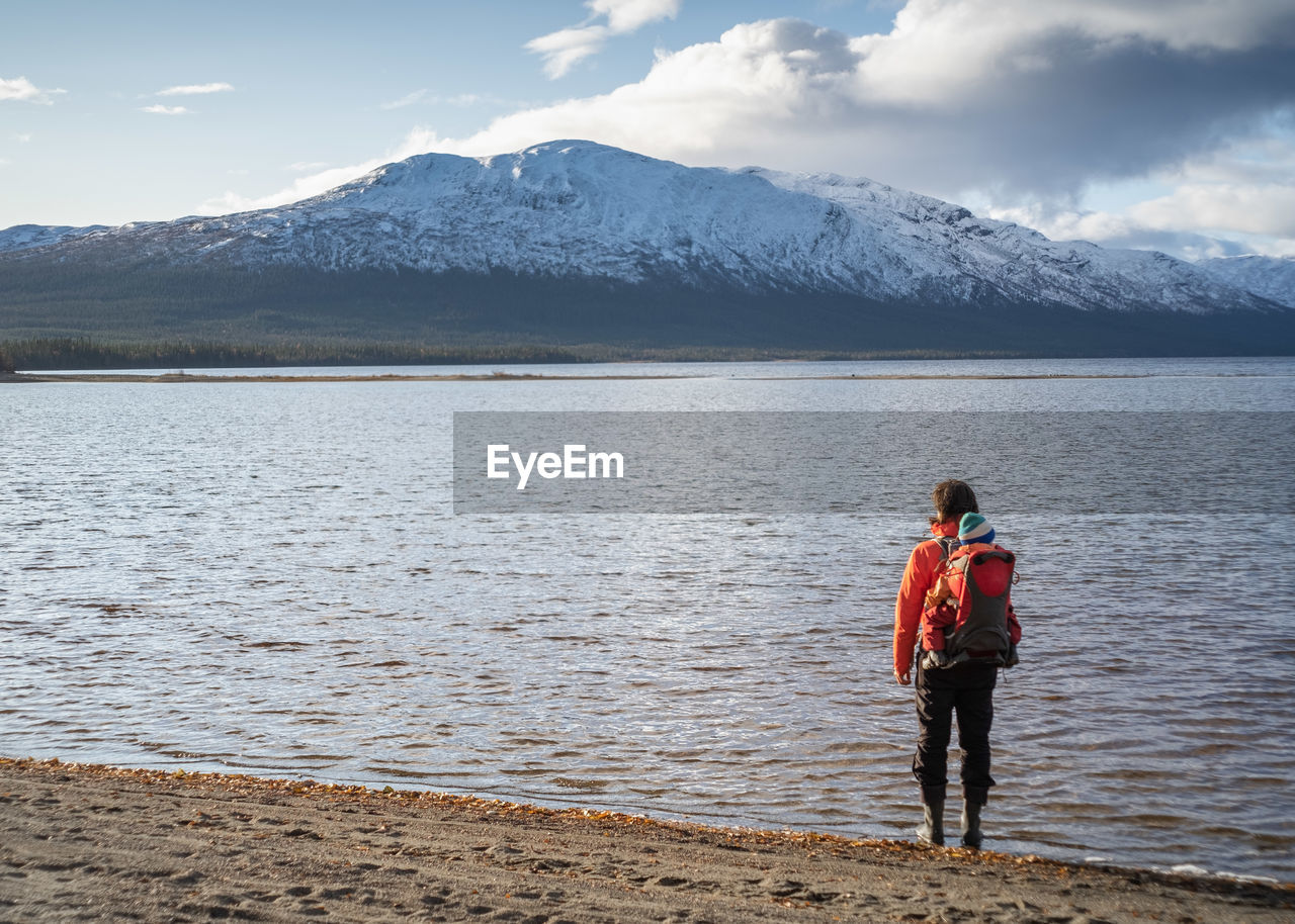 Hiker carrying child in backpack standing at the shore facing a snow capped mountain