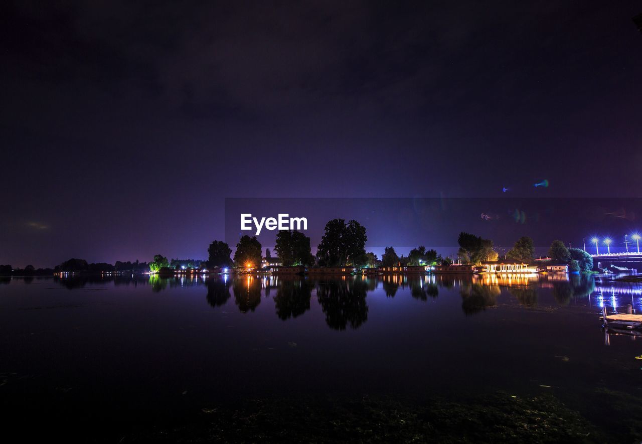 REFLECTION OF ILLUMINATED TREES ON LAKE AGAINST SKY AT NIGHT
