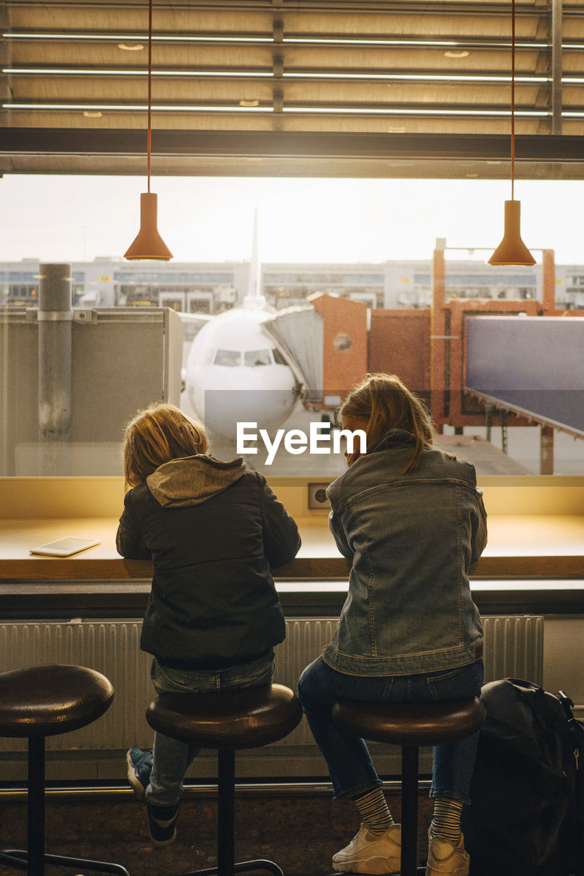 Rear view of brother and sister sitting against window at airport