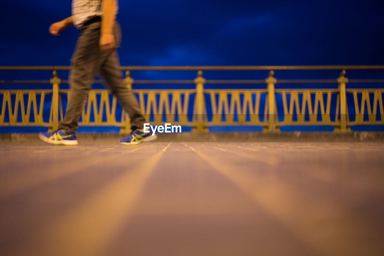 LOW SECTION OF MAN WALKING ON RAILING AGAINST BLURRED BACKGROUND