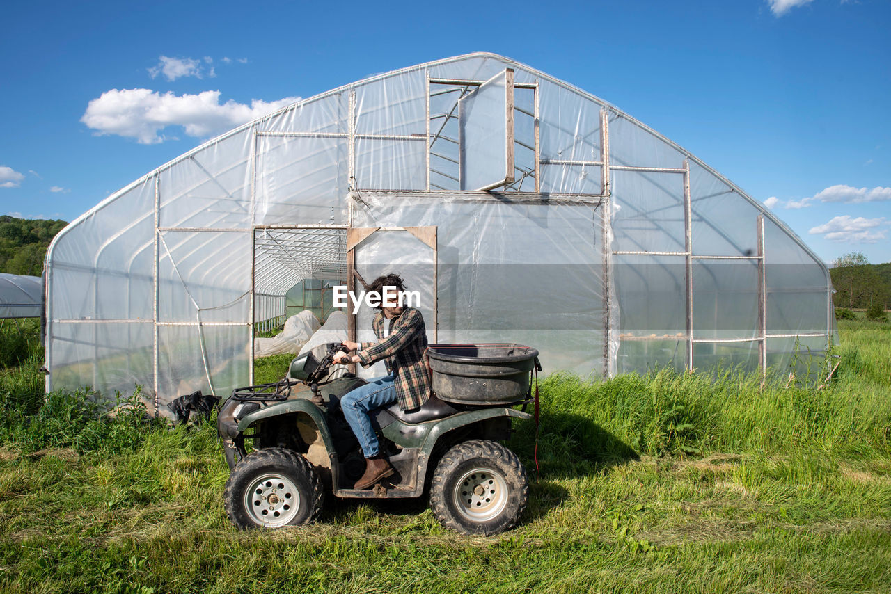 Young farmer on off road vehicle in green sunny field by vegetable greenhouse