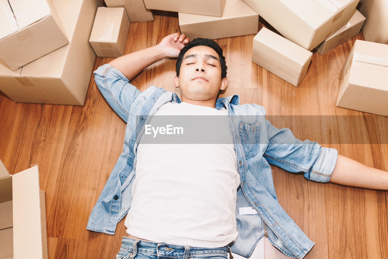 High angle view of young man lying by cardboard boxes on floor at new home