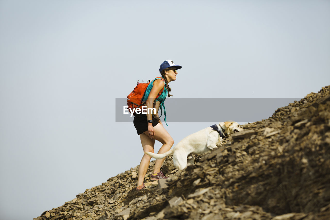 Side view of woman carrying backpack with dog climbing mountain against clear sky