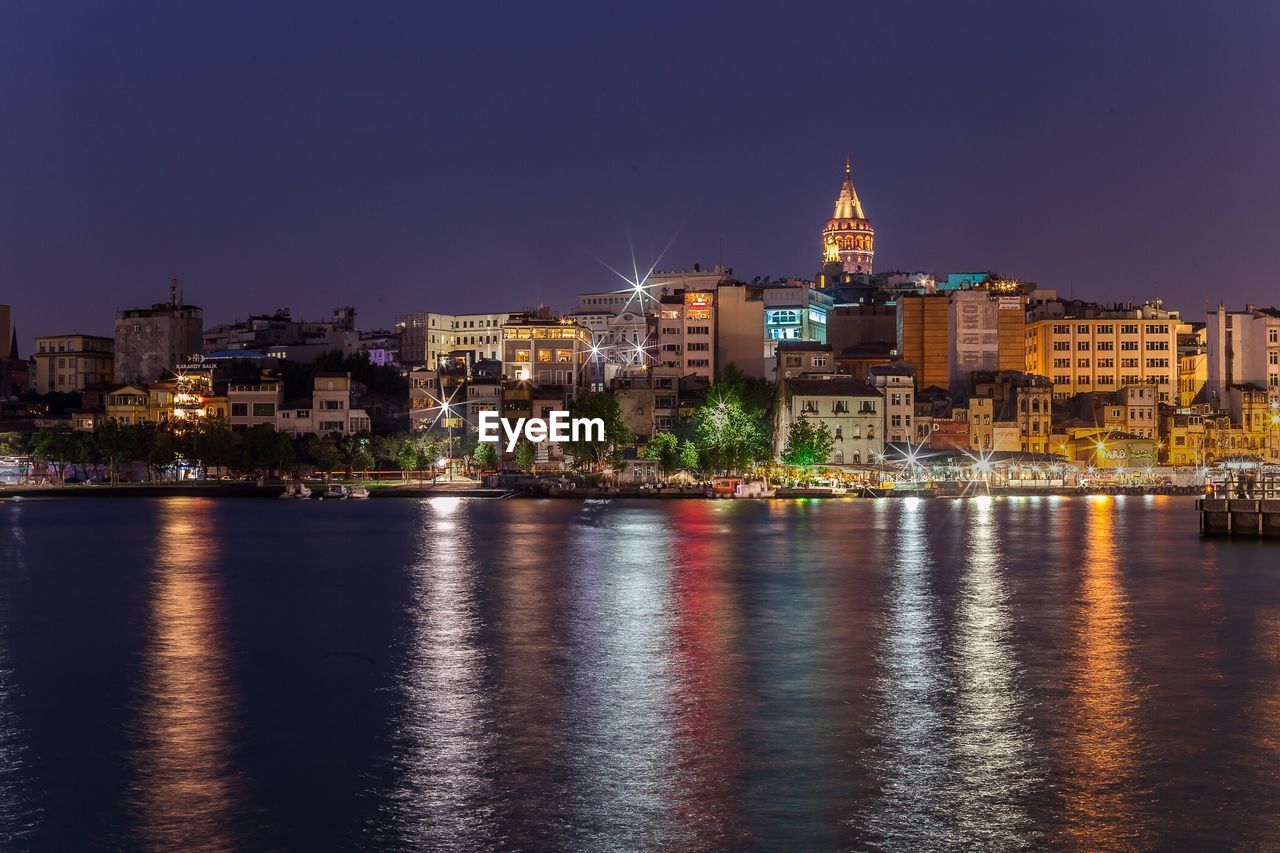 Illuminated galata tower amidst buildings in front of river against clear sky at night