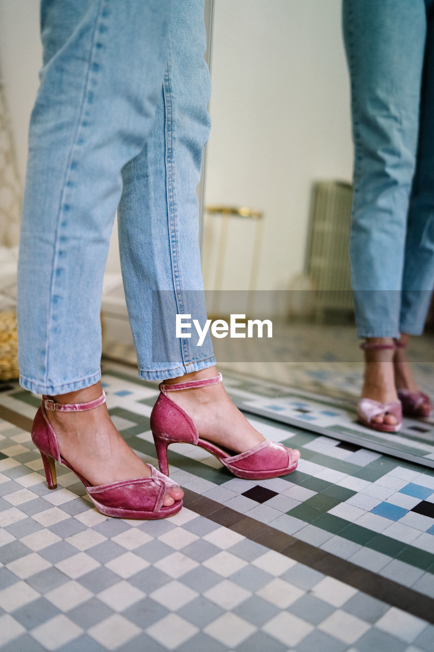Unrecognizable female wearing jeans and open toe heels standing in front of mirror in modern apartment